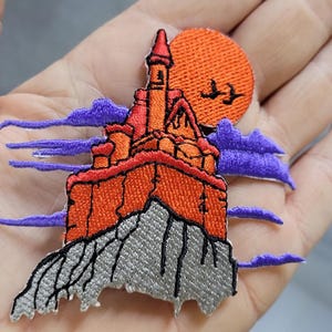 Może przedstawiać: Haftowana naszywka przedstawiająca pomarańczowy zamek na szarym skalistym klifie. Scena zawiera duży pomarańczowy księżyc z dwoma czarnymi ptakami w locie, otoczony poziomymi fioletowymi chmurami na białym tle.