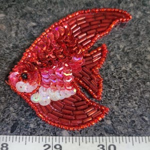 May include: A red sequined fish-shaped applique. The applique is made of red sequins and beads, with iridescent white sequins on the belly. The fish has a black eye and is approximately 3 inches long.