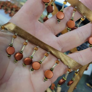 May include: Close-up of decorative trim with a gold satin ribbon base. The trim features hanging strands of round, orange-brown beads and smaller gold beads. The beads are attached with gold and silver accents. The trim is likely used for embellishing clothing or home decor.