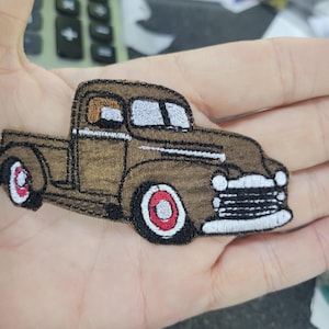May include: Embroidered patch of a vintage brown pickup truck with black tires and red accents. The truck features a detailed design with white stitching outlining the body and windows. This patch is ideal for adding a unique touch to clothing or accessories.