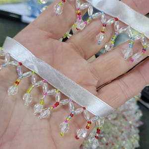 May include: White satin ribbon with beaded fringe. The fringe features clear, iridescent beads and colorful accents of red, yellow, pink, and blue. This decorative trim is suitable for embellishing garments, accessories, or craft projects.