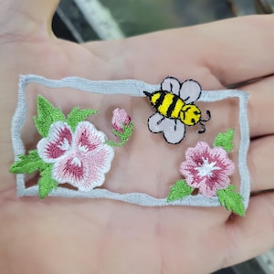 May include: Embroidered patch featuring a bee and pink flowers with green leaves. The bee has yellow and black stripes. The flowers have pink petals with white and red accents. The patch is framed by a light gray border.
