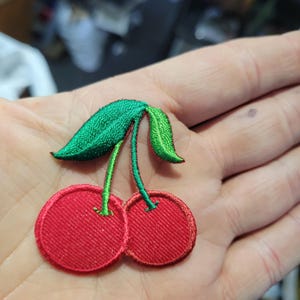 May include: Embroidered cherry patch featuring two red cherries with green stems and leaves. The patch has a detailed, textured design, perfect for adding to clothing or accessories. The patch is a vibrant red and green.