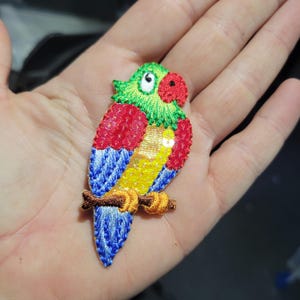 May include: Embroidered parrot patch with a green head, red beak, and colorful wings. The parrot is perched on a brown branch. The wings feature red, blue, and yellow sequins. This decorative patch is suitable for clothing or accessories.