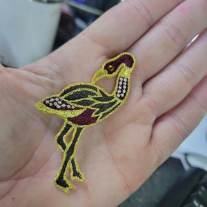 May include: Embroidered patch with a bird design. The patch has a gold outline, black and burgundy details, and a small orange bead for the eye. It is likely designed for application on clothing or accessories.