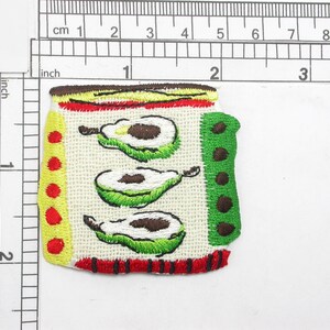 Peut inclure: Un patch brodé avec un fond blanc et trois avocats verts. Les avocats sont brodés avec des pépins bruns et des feuilles vertes. Le patch a une bordure rouge, jaune et verte.