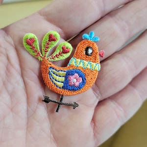 May include: Embroidered bird patch in orange, blue, green, and pink. The bird has a blue crest, a pink flower on its chest, and a decorative pattern around its neck. The patch is embellished with a black arrow.