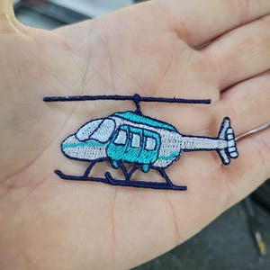 May include: Embroidered patch of a helicopter in shades of blue, white, and navy. The helicopter features a detailed design with a rotor, tail, and landing gear. This patch is suitable for clothing, bags, or other fabric items.