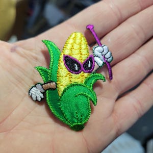May include: Embroidered patch featuring a cartoon corn cob character with purple sunglasses, holding a purple nail. The corn has a yellow center and green husks, with a white glove holding the nail.