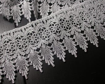 May include: Close-up of silver lace trim with a floral and leaf design. The lace features a repeating pattern of circular floral motifs and delicate, pointed leaf-like edges. The trim is likely used for sewing, crafting, or adding decorative detail to garments.