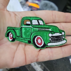 May include: A green embroidered patch of a vintage pickup truck. The truck features a black grill, white headlights, and red and white wheels. The patch is detailed with black outlines and stitching.