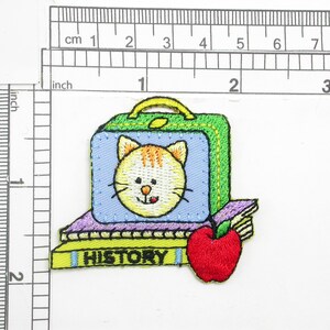 May include: An embroidered iron-on patch featuring a green lunchbox with a cat face on the front. The lunchbox is sitting on top of a stack of books with the word "HISTORY" on the front of the top book. There is a red apple next to the books.