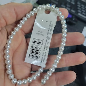 May include: A strand of silver pearls, held in a hand, with a white tag attached. The tag reads "Silver Pearls" and includes a barcode and company information. The pearls are small, round, and a light silver color.