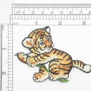 May include: Embroidered iron-on patch of a cute orange and brown tiger cub with black stripes, holding a green branch in its paws. The tiger cub is looking to the right.