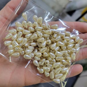 May include: A collection of ivory-colored, teardrop-shaped faux pearls with gold-colored loops. The pearls are contained in a clear plastic bag, and are suitable for jewelry making or craft projects.