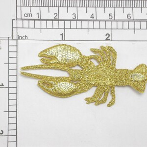 May include: Gold embroidered iron-on patch of a lobster. The lobster is facing right and has a detailed design.