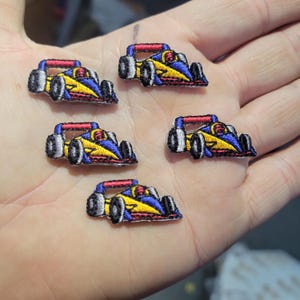 May include: Five embroidered patches of a race car. The patches feature a yellow body, blue accents, and red roll bars. The wheels are black, and the patches are small, suitable for embellishing clothing or accessories.