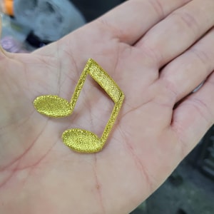 May include: A gold-coloured musical note-shaped patch. The patch is made of a textured, woven material. The musical note is a double quaver, with two connected oval-shaped notes and a vertical line.