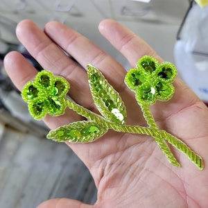 May include: A beaded floral applique in shades of green. The applique features two flowers and several leaves, all embellished with sequins and beads. The stems are a lighter green, and the flowers are a brighter, more vibrant green.