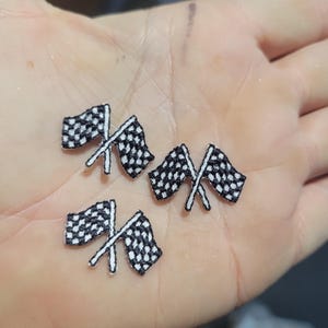 Pode incluir: Pequenas bandeiras de corrida quadriculadas bordadas em preto e branco. As bandeiras são cruzadas e têm mastros brancos. Estes patches são ideais para adicionar um tema de corrida a roupas ou acessórios.