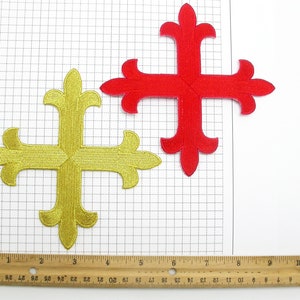 Peut inclure: Deux patchs brodés thermocollants en forme de croix avec un motif de fleur de lys. La croix de gauche est dorée et la croix de droite est rouge.