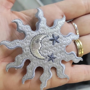 May include: A silver embroidered patch in the shape of a sun with wavy rays. The centre features a crescent moon with a face and two small dark grey stars. The embroidery has a textured, metallic sheen throughout the entire sun design.