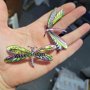 May include: Two embroidered dragonfly patches. The dragonflies have detailed wings with green, pink, and white patterns. The bodies are a light purple colour. The patches are held in an open hand.