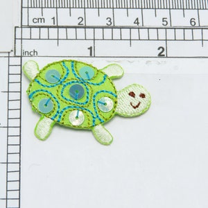 Peut inclure: Applique de tortue brodée verte et bleue avec des paillettes. La tortue a un visage souriant et mesure environ 5 cm de large.