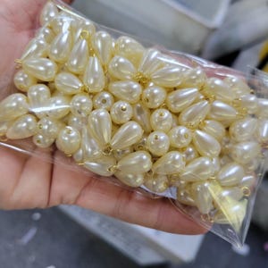 May include: A clear plastic bag filled with ivory-colored faux pearl beads. The beads are a mix of round and teardrop shapes, with small gold-colored loops for stringing. The pearls have a glossy finish and are suitable for jewelry making and crafts.