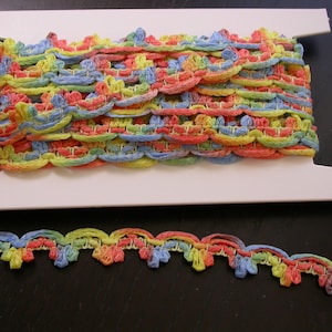 Op de afbeelding: Een spoel met kleurrijk gecrochete trim met een gegolfde rand. De trim is gemaakt van zacht, getextureerd garen in tinten blauw, geel, roze en groen.