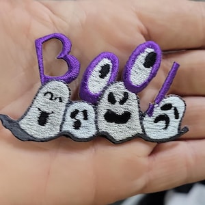 May include: An embroidered patch featuring the word Boo! in purple thread, with the letters integrated into four white ghost figures. Each ghost has a distinct black stitched facial expression, including smiling, surprised, and frowning faces.