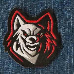 May include: Black and white wolf's head embroidered patch with a red outline. The wolf has a fierce expression with sharp teeth and a snarling mouth.