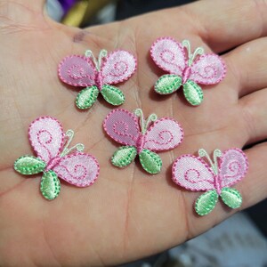 May include: Five pink butterfly appliques with green accents are displayed. Each butterfly has a detailed design with a pink body, pink wings, and green leaves. The appliques are likely for crafts or embellishments.