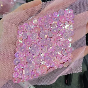 Pode incluir: Close-up de um pedaço de tecido de tule rosa adornado com lantejoulas redondas e iridescentes. As lantejoulas são densamente embaladas em um padrão quadrado, criando um efeito brilhante. O tule tem uma textura de malha fina, e as lantejoulas refletem várias tonalidades de rosa e roxo.