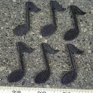 May include: Six black embroidered musical note patches arranged in two rows. A measuring tape is visible at the bottom, showing the scale in inches with numbers 28, 29, and 30 clearly printed on the white tape against the dark textured surface.