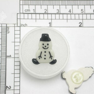 May include: White plastic button with a black snowman design. The snowman has a black hat, eyes, mouth, and buttons. The button is approximately 1 inch in diameter.
