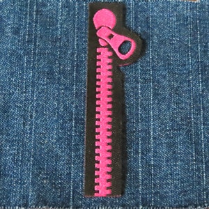 May include: A pink and black zipper patch on a blue denim background. The zipper is a bright pink color with a black outline. The denim fabric has a classic blue wash.