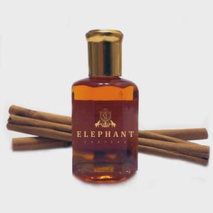May include: A rectangular glass bottle with a gold cap, filled with a golden-brown liquid. The label reads "ELEPHANT CHATEAU" with a crest. Cinnamon sticks are arranged around the bottle. The overall aesthetic is warm and inviting.