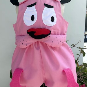 May include: A pink sleeveless romper featuring a character face with large white eyes, black eyebrows, and a red mouth. The garment includes black ear-like accents at the shoulders, a snout section with small black markings, and two pink felt feet.