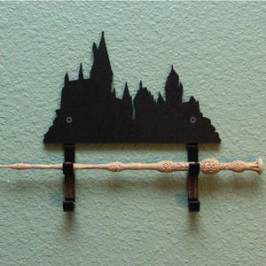 May include: A black metal silhouette of Hogwarts castle with two hooks on the bottom. A light brown wand is hanging from the hooks.