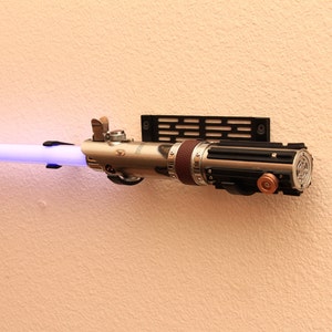 Könnte beinhalten: Ein silbernes und schwarzes Lichtschwert mit einer violetten Klinge ist an einer Wand mit einer schwarzen Metallhalterung befestigt. Das Lichtschwert hat einen braunen Ledergriff und einen silbernen Metallknauf.