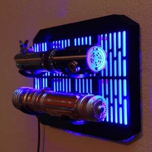 May include: A black wall mount with a blue light behind it. The wall mount has a grid pattern and two lightsaber holders. The lightsabers are silver and orange with black handles.