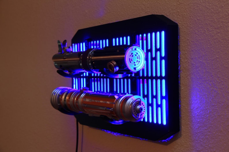 May include: A black wall mount with a blue light behind it. The wall mount has a grid pattern and two lightsaber holders. The lightsabers are silver and orange with black handles.