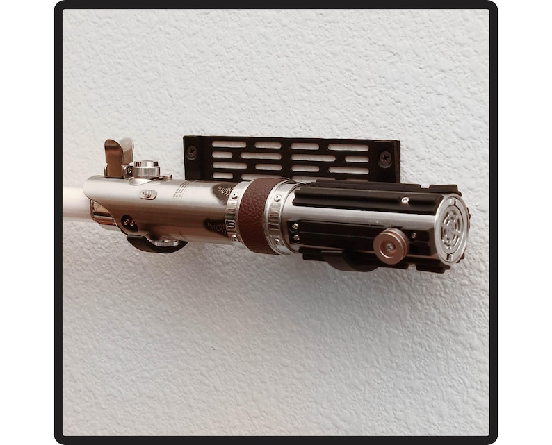 K&ouml;nnte beinhalten: Eine silberne und schwarze Lichtschwert-Replik, die an einer Wand mit einer schwarzen Wandhalterung aus Metall befestigt ist. Das Lichtschwert hat einen braunen Ledergriff und eine silberne Klinge.