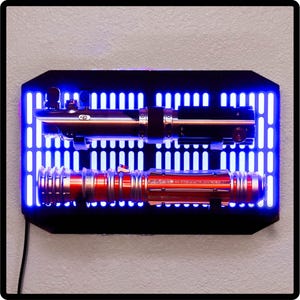 May include: Two lightsabers mounted on a black wall mount with a blue light grid background. The top lightsaber is black and silver, the bottom lightsaber is red and silver.