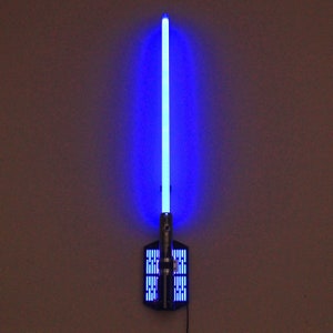 May include: A blue lightsaber wall mount with a black base and a silver metal plate with a grid pattern. The lightsaber is lit up and the blade is a bright blue color.