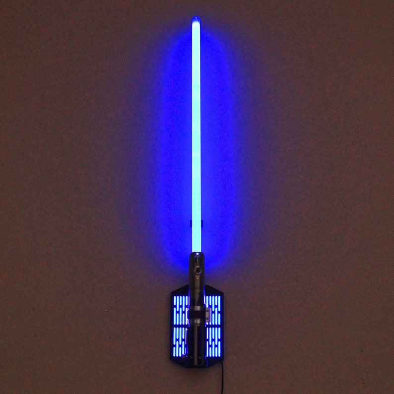 May include: A blue lightsaber wall mount with a black base and a silver metal plate with a grid pattern. The lightsaber is lit up and the blade is a bright blue color.