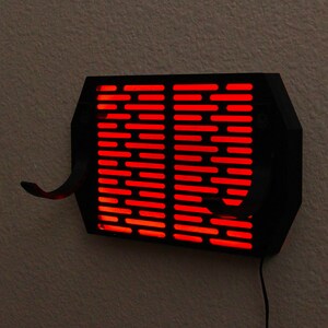Puede incluir: Una l&aacute;mpara de pared negra con forma rectangular y una serie de ranuras horizontales. La l&aacute;mpara emite una luz roja brillante. Dos ganchos curvos se extienden desde la parte inferior. Se ve un cable de alimentaci&oacute;n negro.