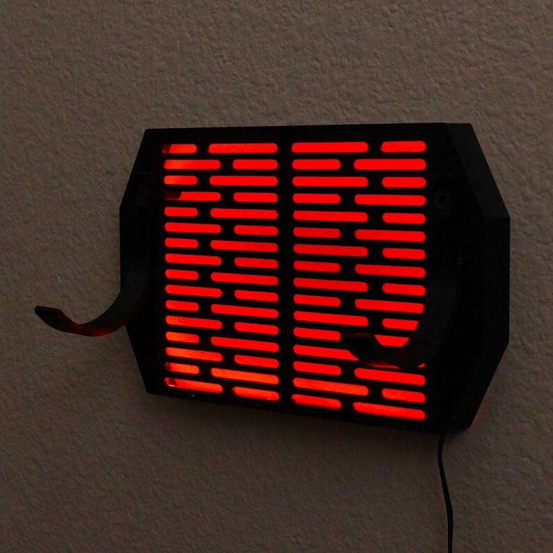 Puede incluir: Una l&aacute;mpara de pared negra con forma rectangular y una serie de ranuras horizontales. La l&aacute;mpara emite una luz roja brillante. Dos ganchos curvos se extienden desde la parte inferior. Se ve un cable de alimentaci&oacute;n negro.
