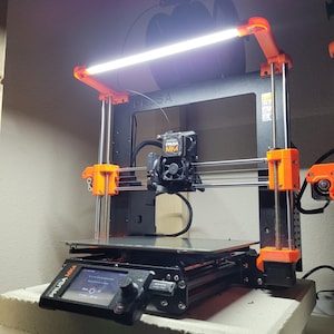 May include: A black 3D printer with orange accents. The printer has a white LED light bar attached to the top. The printer has a small LCD screen on the front. The text "PRUSA MK4" is visible on the printer.